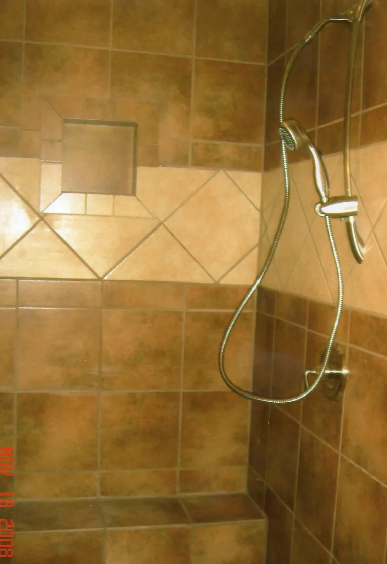 Newly remodeled bathroom with glossy brown tiles, built-in corner soap shelf, and a silver handheld shower head, reflecting a modern and functional design.