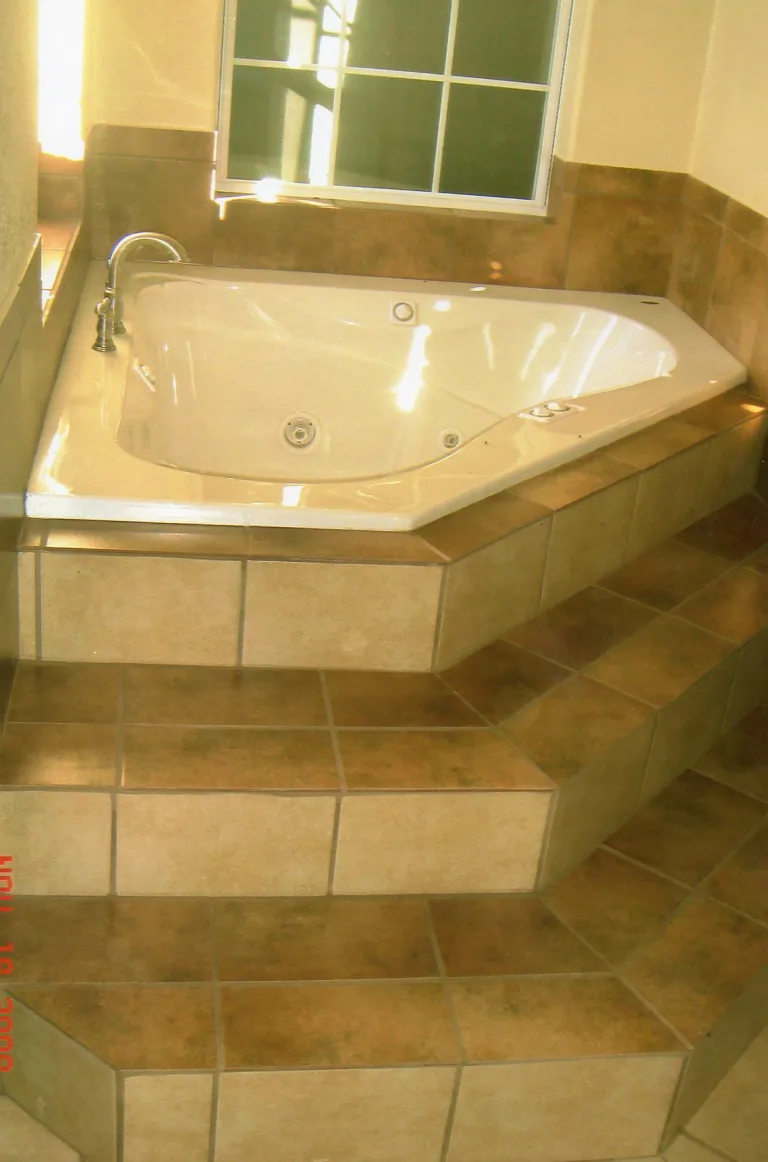 Spacious bathroom corner with a large jacuzzi bathtub on raised beige tiling, featuring built-in steps and a bright window with grid design, reflecting a luxurious and inviting ambiance.