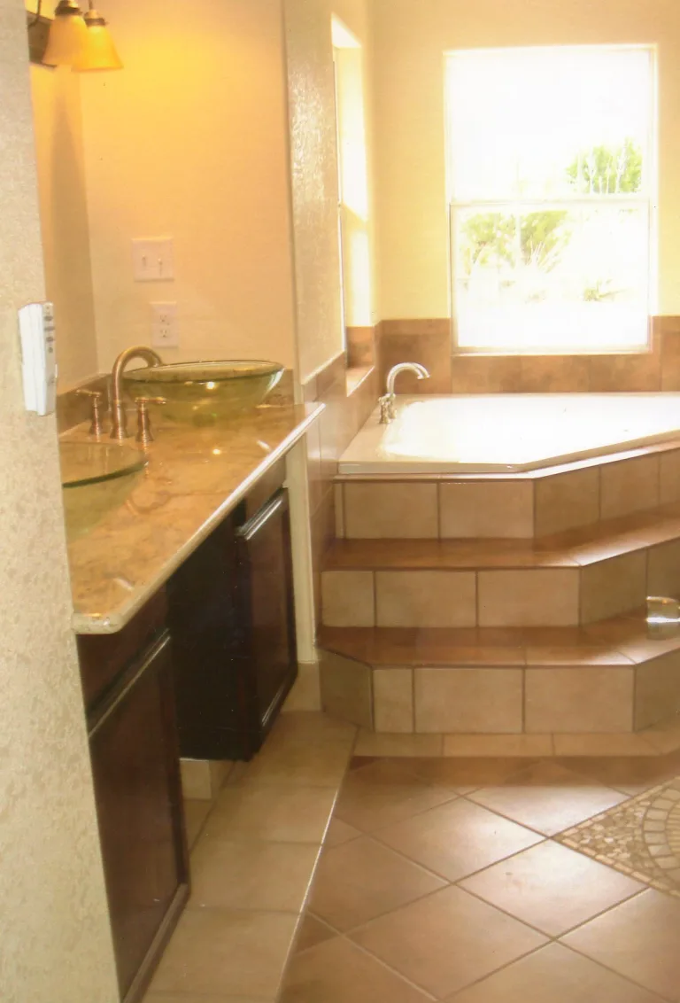 Elegant bathroom interior featuring a corner jacuzzi bathtub with beige tile steps, a granite countertop with a vessel sink, and a large window with a view, creating a bright and sophisticated space.