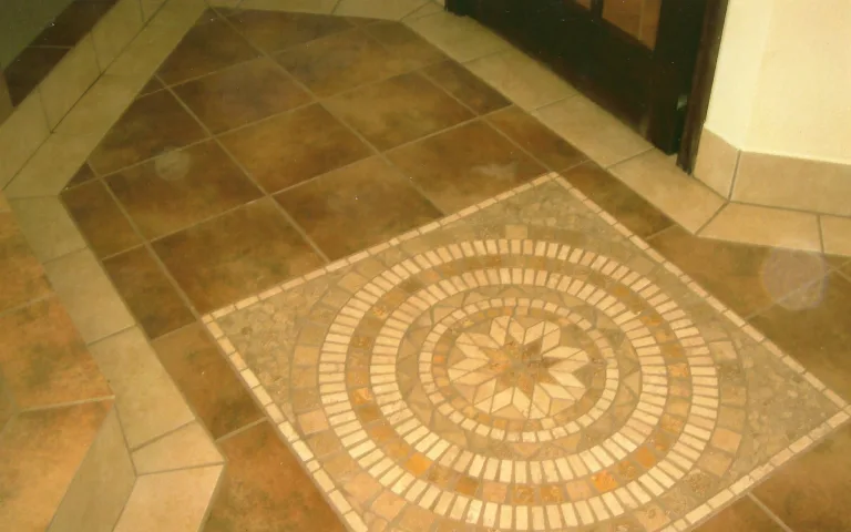 Detailed view of a bathroom floor with a decorative circular mosaic tile centerpiece, surrounded by large beige tiles, part of a stylish bathroom remodel.