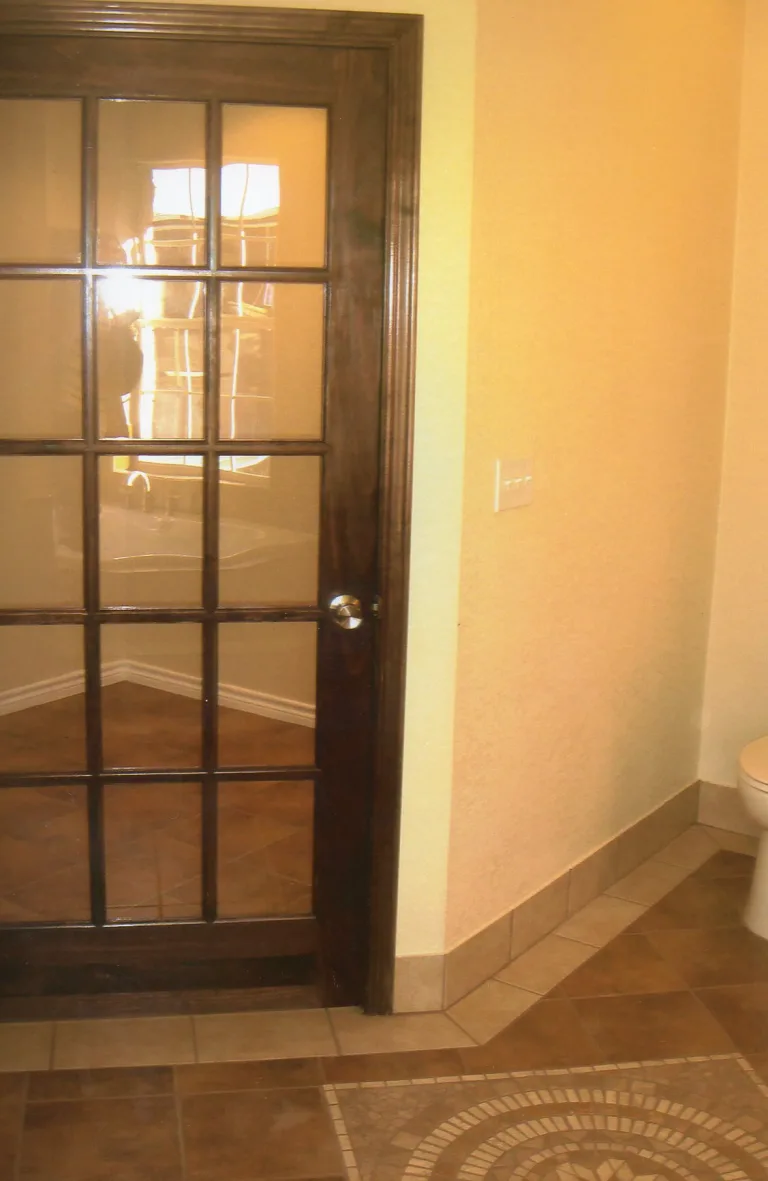 Modern bathroom entry with dark wooden door featuring frosted glass panes, tiled flooring with mosaic details, and a glimpse of a white toilet, part of a comprehensive bathroom renovation.