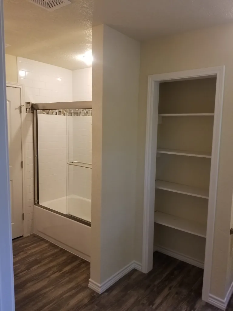 Modern bathroom design featuring wood-look tile flooring, a white bathtub with subway tiles and mosaic trim, and a built-in white shelving unit for storage.