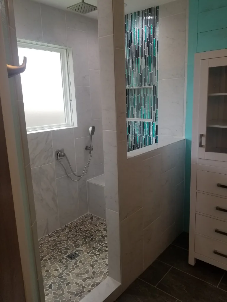 Luxurious bathroom shower with marble wall tiles, decorative vertical mosaic tile accent, pebble floor, handheld showerhead, and a window for natural light.