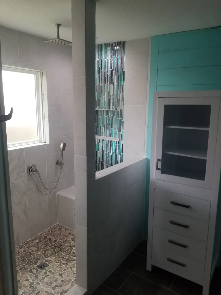 Spacious shower with pebble tile flooring, a vertical strip of colorful mosaic tiles, a large rainfall showerhead, and a modern white storage cabinet against a turquoise wall.