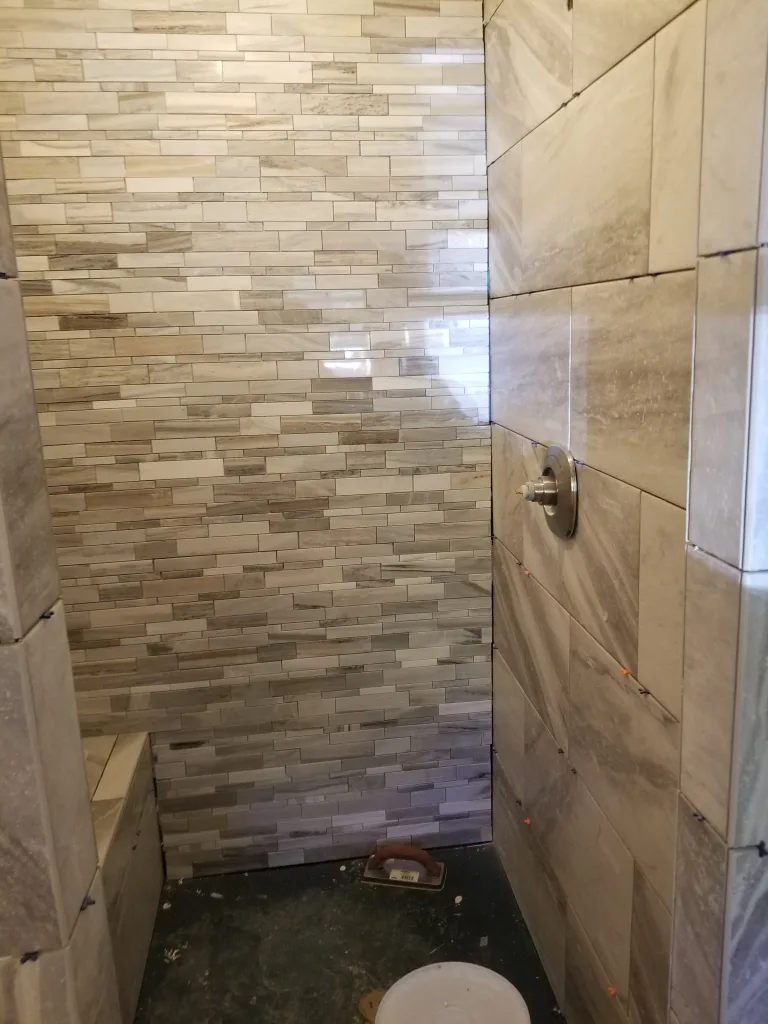 Bathroom renovation in progress with a shower featuring multi-toned gray strip mosaic tiles, a corner seat, and construction tools visible, indicating work underway.