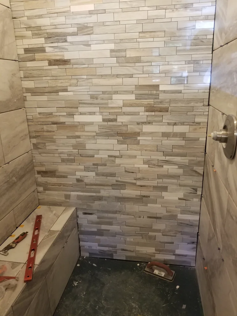 Under construction shower area with horizontal multi-shade mosaic tiles and a built-in bench, including a level and trowel on the floor indicating ongoing work.