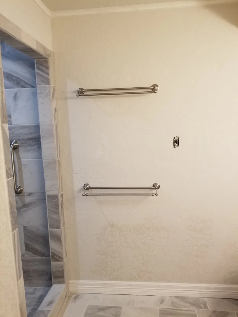 Neutral-toned bathroom wall with modern brushed nickel towel bars and a glimpse into a shower with marble tiling, illustrating a clean and functional design.