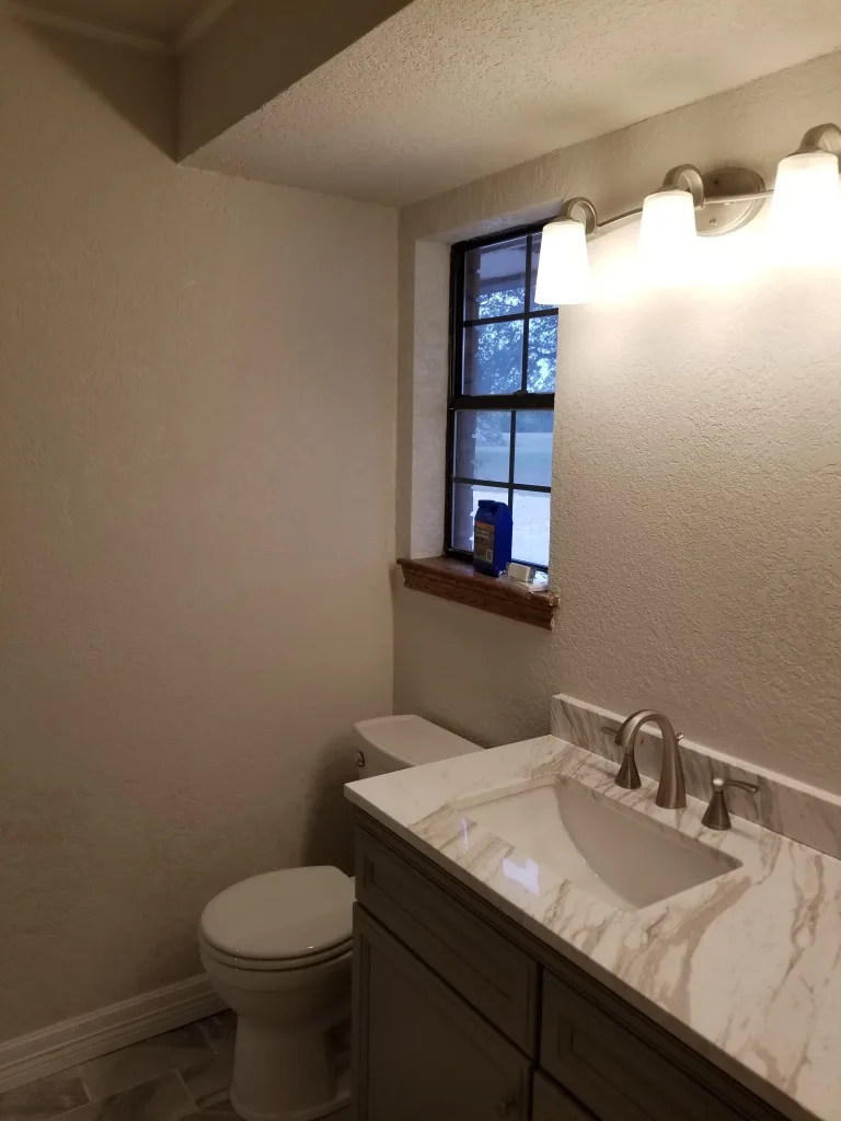 Bright bathroom with a window allowing natural light, featuring a vanity with a marble countertop and dual sinks, next to a white toilet under a wall-mounted light fixture.