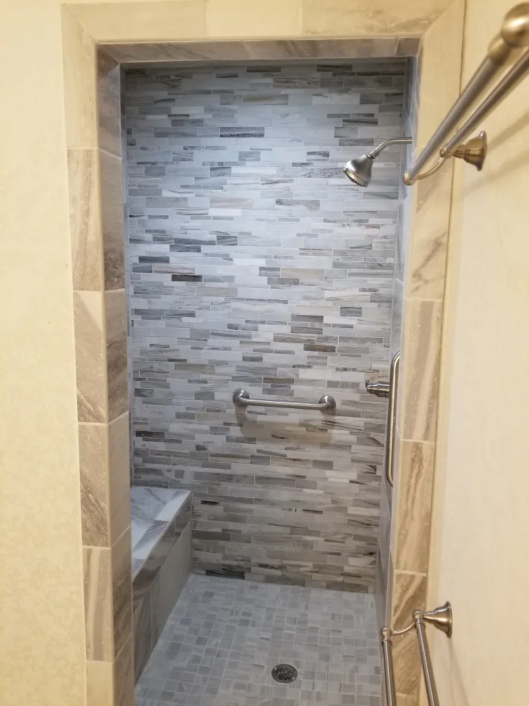 Sleek and stylish shower with a mix of elongated mosaic tiles in shades of gray, built-in safety grab bars, and a modern shower head, all enhancing the shower's contemporary design.