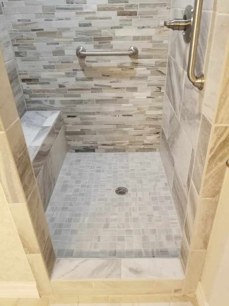 Modern shower featuring a mix of horizontal mosaic tiles in neutral tones, a convenient corner bench, and safety grab bars, with a square tile floor design.