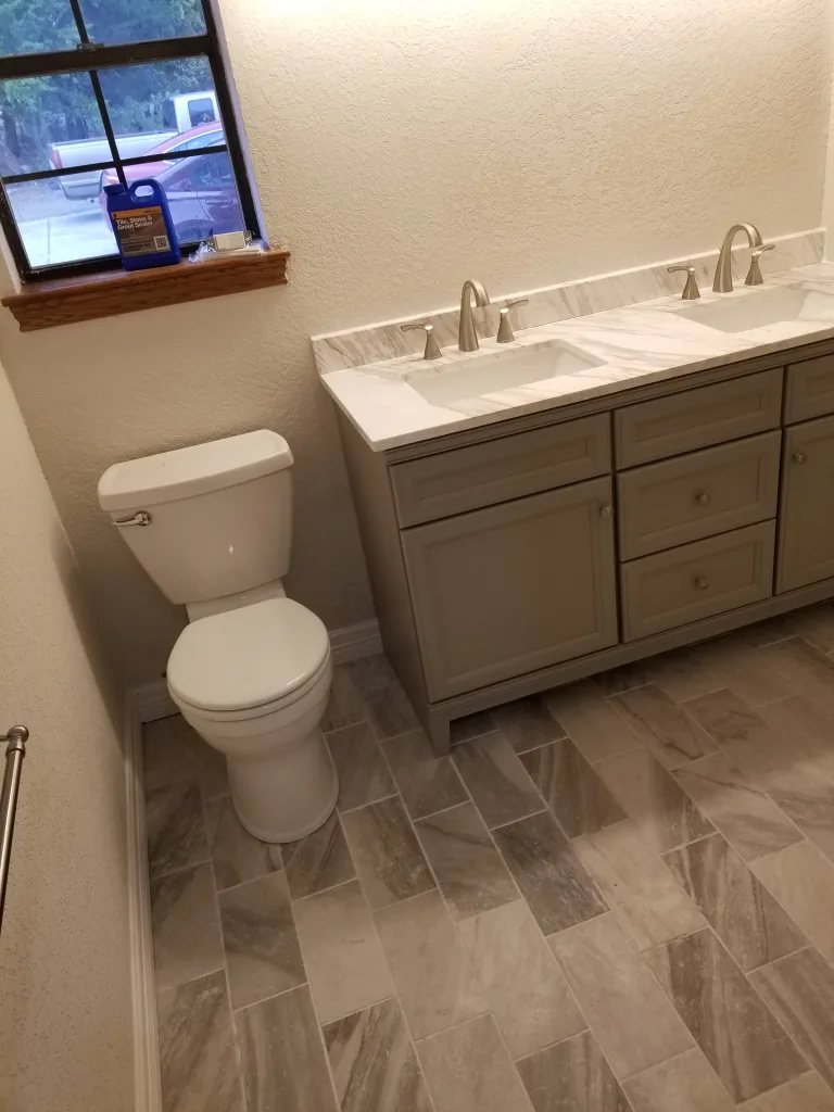 Spacious bathroom with a white dual-sink vanity, marble countertop, and a window providing natural light, complemented by a herringbone pattern tiled floor.