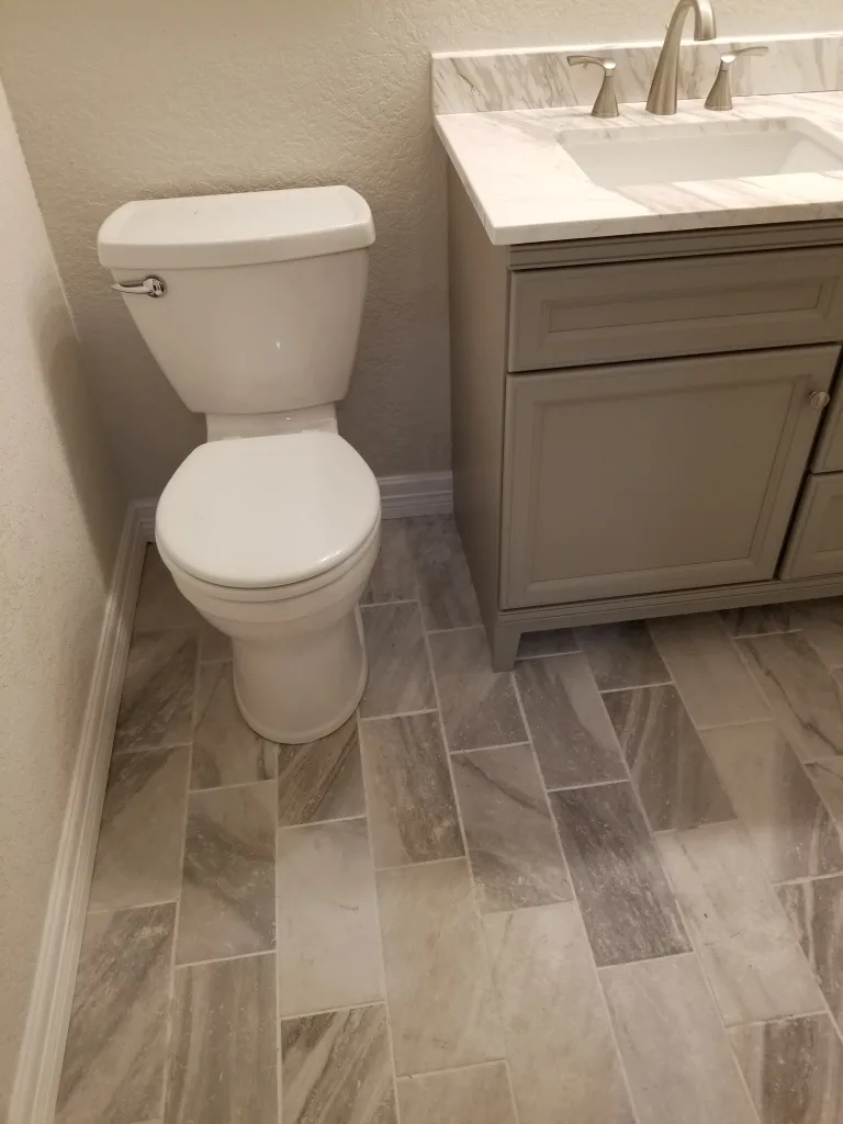 Neat bathroom corner with a classic white toilet and a gray vanity cabinet with a marble top and undermount sink, set against a herringbone tile floor.