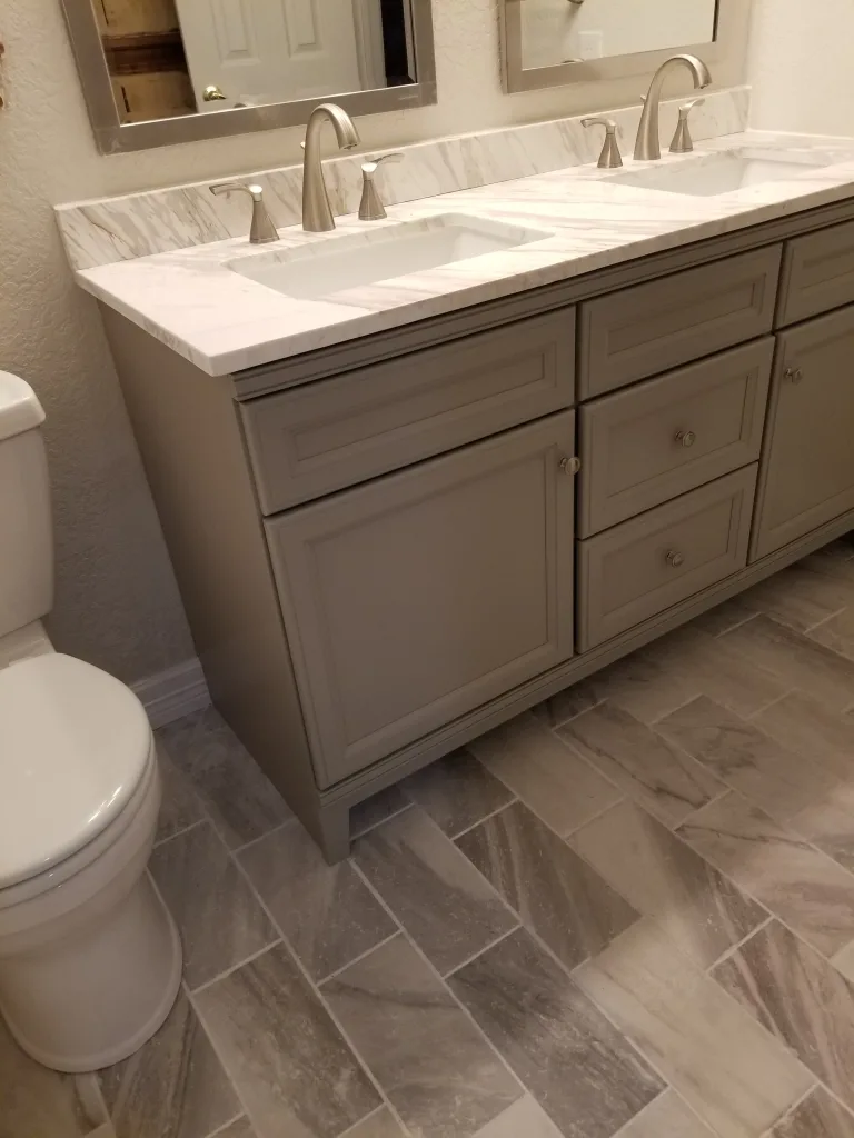 Refined bathroom with a large double-sink vanity, marble countertop, brushed nickel faucets, and herringbone pattern tile floor next to a classic white toilet.