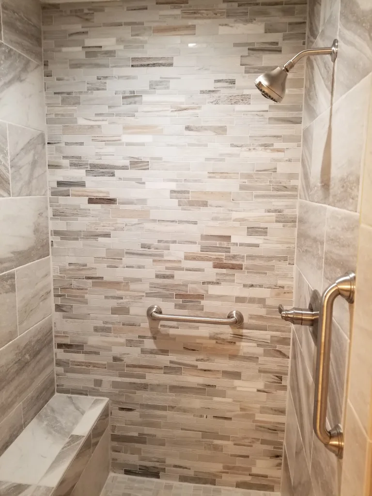 Elegant shower space with varied shades of beige and gray linear mosaic tiles, a built-in corner shelf, safety grab bar, and a modern rainfall showerhead.