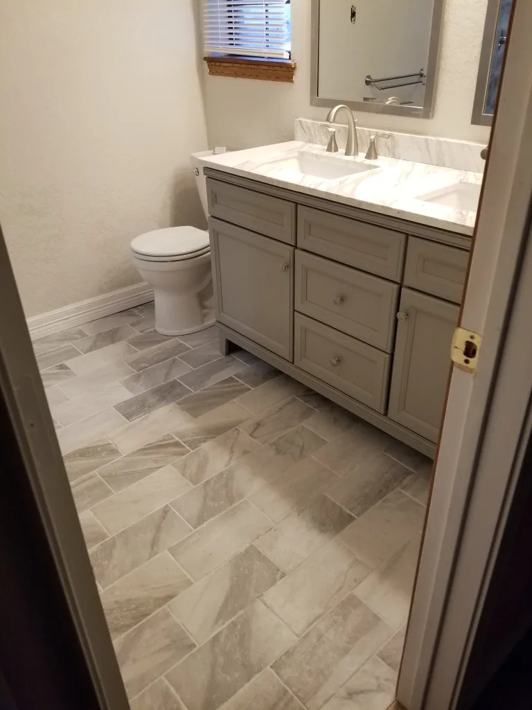 Elegant bathroom with herringbone pattern tile flooring, classic white vanity with marble top, and a white toilet beside a window with wooden blinds.