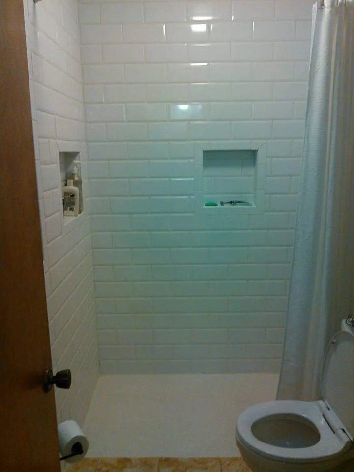 Bathroom interior with white subway tile shower, built-in wall shelf for bath products, white shower curtain, and adjacent toilet with toilet paper holder.