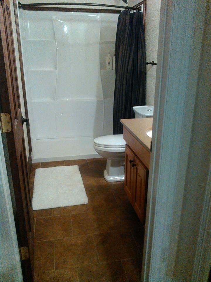 Compact and functional bathroom with a white shower stall, classic white toilet, wooden vanity with beige countertop, and a plush white mat on a tiled floor.